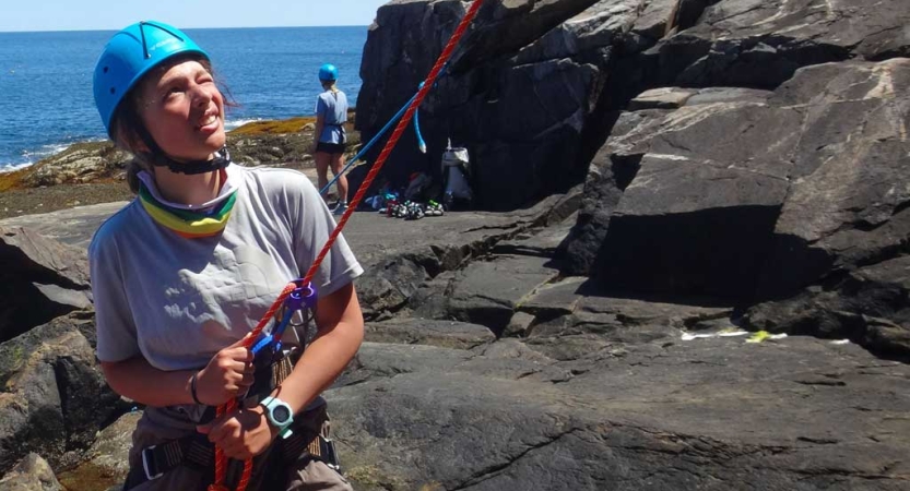 A student wearing safety gear looks up as they belay a rock climber